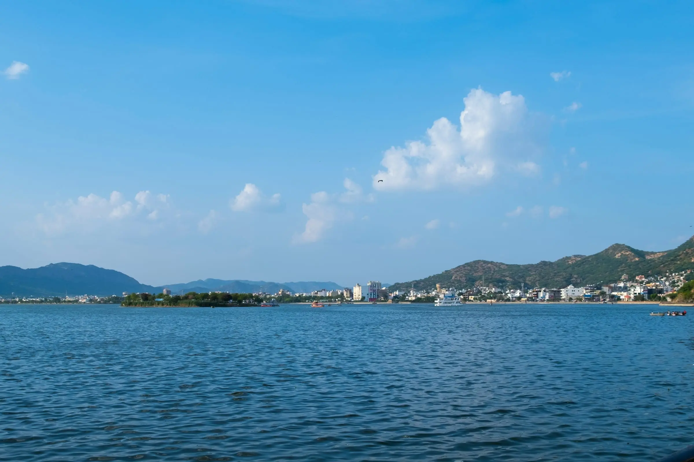A wide lake in Ajmer under a blue sky, with hills on the far shore